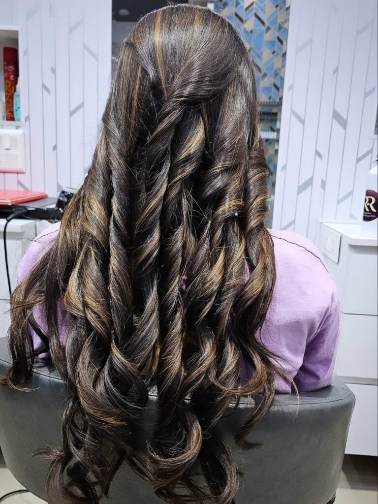 "Back view of a client with long, glossy dark brown hair styled in soft, defined curls with a half-up twist. The elegant hairstyle showcases smooth texture, subtle highlights, and a polished salon finish in a modern beauty salon."